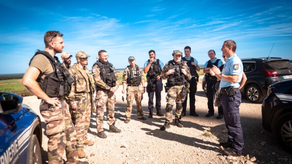 Un gendarme debout entouré d'autres gendarmes et de militaires de l'armée de Terre s'exprime devant tous. Ils sont dehors, entourés de véhicules. derrière eux, on aperçoit un champ. Le ciel est bleu.