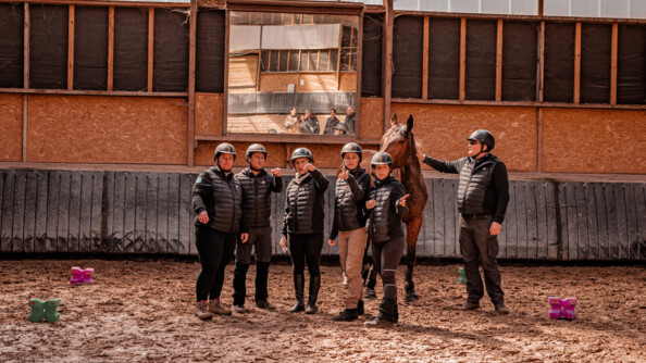 Les six participants au stage Cent'Or, pied à terre, dans le manège du Centre d’instruction du régiment de cavalerie de la Garde républicaine, à Saint-Germain-en-Laye. Cinq pointent quelque chose du doigt. Le sixième à droite en arrière plan carresse un cheval