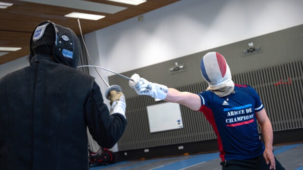Maxime Pauty, vêtu du survêtement bleu et rouge de l'armée de champions, touche son maître d'arme, de dos, vêtu de noir, qui tente une parade
