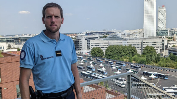 Un gendarme en buste sur les hauteurs devant des immeubles blancs. Au sol de nombreux bus