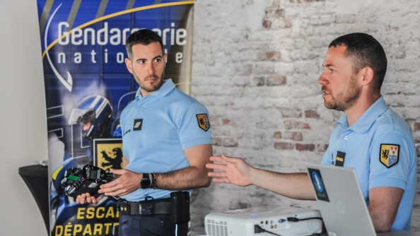 Deux gendarmes debout dans une salle en train d'expliquer le fonctionnement de leur matériel de relevé de vitesse. Au fond à gauche un kakémono de l'escadron départemental de sécurité routière du 59. Le gendarme de droite est derrière une petite table haute équipée d'un ordinateur et projecteur; celui de gauche l'écoute et tient en main l'appareil de mesure
