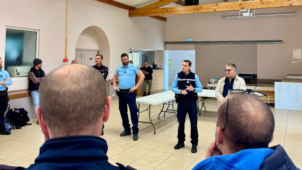un commandant de gendarmerie entouré de gendarmes et de civils dans une salle donne un briefing