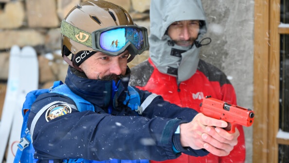 un gendarme coiffé d'un casque de ski pointe un pstolet rouge d'entraînement. en arrière-plan à droite, un homme en rouge