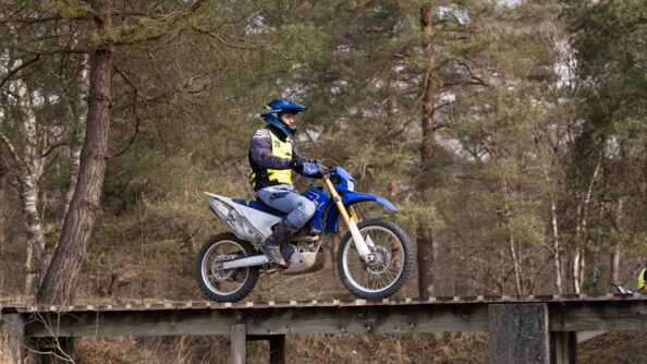 Un motard de la gendarmerie sur une moto cross bleue passe sur un pont de bois dans une forêt de résineux