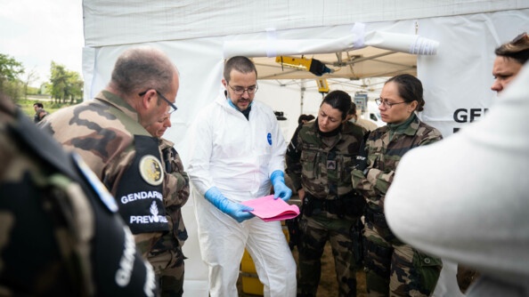 À l'entrée d'une tente, un scientifique en combinaison blanche fait un compte rendu des données relevé à un groupe de gendarmes en treillis regrouper autour de lui.