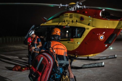 Un gendarme, dos à l'objectif, marche vers un hélicoptère posé sur le tarmac, de nuit. Dans le dos de sa chasuble orage est écrit : "Secours en montage - Gendarmerie".