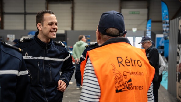 Un gendarme souriant échange avec un homme portant une veste sans manches orange fluo portant la mention "retro passion Rennes"