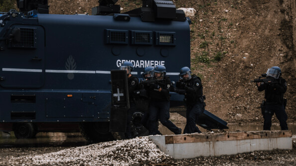 Cinq gendarmes mobiles en colonne d'assaut protégés par un bouclier et équipés d'armes longues remontent le long du Centaure