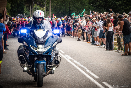 Un motocycliste de la gendarmerie ouvre la route d’une course cycliste féminine, suivi de près par un peloton de coureuses. La moto bleue est équipée de gyrophares allumés. De nombreux spectateurs bordent la route des deux côtés, applaudissant et filmant les cyclistes. L’ambiance est festive et animée.