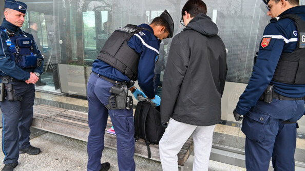 3 gendarmes sont entrain de fouiller le sac &agrave; dos d'un jeune homme sous un abri de train.