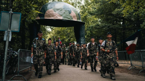 Un groupe de militaires, en tenue kakie, avance face à l'objectif. Ils passent sous un auvent représentant un casque kaki.