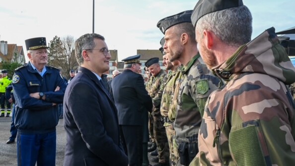 Gérald Darmanin se tient debout, de biais, face à des militaires en tenue kakie. Il regarde l'un d'eux dans les yeux. Derrière le ministre se tient le directeur général de la gendarmerie nationale, en tenue bleue marine, portant un képi et tenant ses bras croisés contre sa poitrine.