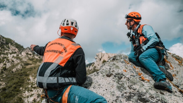 Deux gendarmes sur un rocher, portant une chasuble "secours en montagne", regardant vers les hauteurs