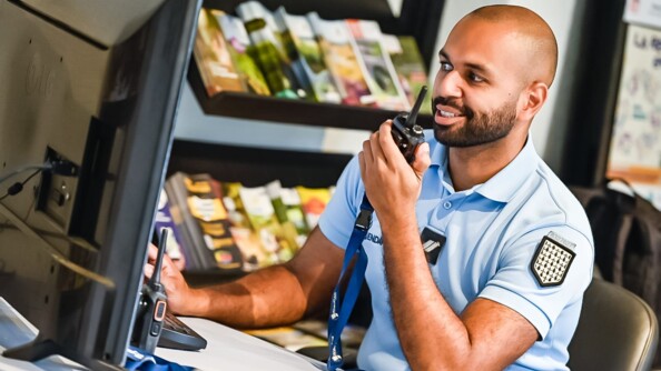 Assis derrière un bureau , souriant, un gendarme tient un talkie-walkie de sa main gauche. Devant lui, un écran d'ordinateur et en arrière-plan un étagère avec des prospectus