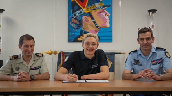Assis derrière une table, un para-sportif de la haut-niveau de la gendarmerie (SHND-G) souriant et s'apprêtant à signer son contrat de SHND. Sa main gauche est atrophiée. A droite, un général de gendarmerie, et à gauche, un commandant de l'armée de terre
