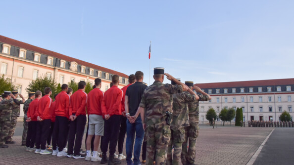 Au premier plan, sur la place d'armes, vue de dos des sportifs en tenue de sport et des militaires de la gendarmerie en treillis kaki, au garde-à-vous. De l'autre côté de la place d'armes, une compagnie d'élèves gendarmes au garde-à-vous. Au fond et à gauche, les bâtiments de l'école.