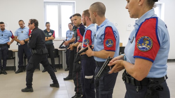 Des élèves en ligne, pistolet à la main regarde l'instructeur qui montre une position de tir