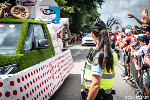 Une gendarme en uniforme, avec un gilet tactique jaune fluo et une queue de cheval, vue de profil, est positionnée sur le bord de la route, tandis que les véhicules de la caravane du Tour de France passent devant elle. De l'autre côté de la chaussée et derrière elle, derrière des barrières métalliques, sont massées de nombreuses personnes qui applaudissent et agitent des drapeaux dans une ambiance festive.