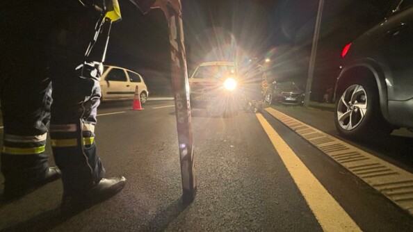Les jambes d'un gendarme tenant un stop stick, de nuit face &agrave; une voiture aux phares allum&eacute;s.