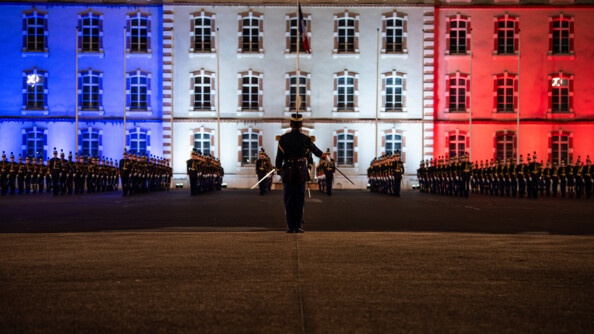 Un officier de l'EOGN, de dos, au centre, salue au sabre les deux promotions d'élèves de l'EOGN, alignés devant l'état-major de l'Ecole, illuminé de bleu blanc et rouge