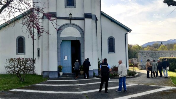 Un gendarme de dos discute avec un homme à sa droite. A droite un groupe de personne agées discutent. Un parvis goudronné mène à une petit église romane blanche à porte d'entrée bleue. En fond de paysage des arbres et des collines sous un ciel gris