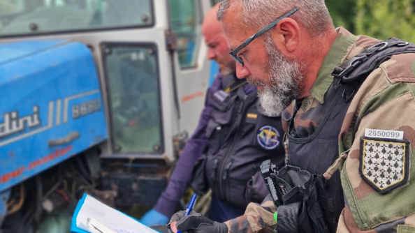Deux gendarmes, accroupis à côté d'un tracteur, relèvent des informations. L'un d'eux prend des notes sur un calpin.