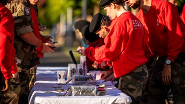 de part et d'utres d'une table recouverte de goodies, des hommes et femmes en treillis avec un sweat rouge et un béret noir