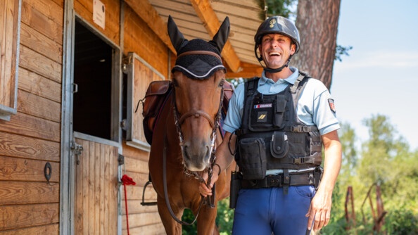 Devant le box, le cheval à gauche, le cavalier à droite, tenant les rênes sous la gorge du cheval. Debout, tout équipé avec son gilet pare-balle et sa bombe d'équitation, il rit aux éclats