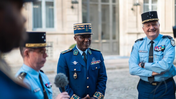 Dans une cour extérieure pavée, se tiennent de bout trois hommes. Deux autorités de la gendarmerie nationale, entre lesquelles se tient le commandant de la gendarmerie congolaise. Sur la gauche, l'homme tient un micro. En arrière plan, on aperçoit la porte et les fenêtres d'un bâtiment, ainsi que des lanternes.t