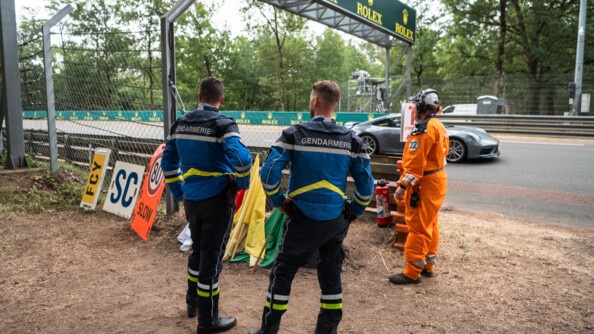 Deux gendarmes sont avec un commissaire de piste et ils discutent. Derrière une porshe passe sur le circuit.