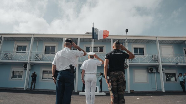 Deux gendarmes et un préfet de dos saluant le drapuea français devant un bâtiment à la façade bleu ciel et blanc