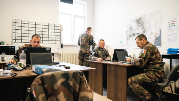 Dans une salle aux murs blancs, quatre bureaux se font face, deux par deux, avec des ordinateurs portables et du matériel de bureau. Les deux bureaux de droite et celui du fond à gauche sont occupés par des prévôts, travaillant. Au fond à droite, derrière son camarade, un prévôt de dos, debout, en train de faire couler le café. Le mur de droite arbore une carte de la Roumanie et de la documentation, le mur du fond un tableau blanc magnétique du service mensuel