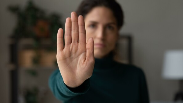Une femme brune vêtue d'un pull vert sombre, se tient debout au centre de l'image. Devant elle, à hauteur d'épaule, elle tend sa main droite, comme pour dire "stop". La femme se trouve dans ce qui semble être un salon. Le second plan de l'image est flouté, mais on devine une lampe sur pied avec un abajour blanc un meuble.
