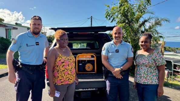 Deux gendarmes posent, souriants, autour de deux femmes réunionnaises, qui sourient également, après le passage du cyclone Garance à La Réunion. Le temps ets beau et le ciel est bleu. Les quatre personnes s etrouvent debout dans la rue, devant un véhicule dont le coffre est ouvert, et dévoile un dérouleur de câble.