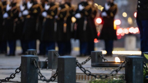 Gros plan sur la flamme du Soldat inconnu, prot&eacute;g&eacute;e par des plots et des cha&icirc;nes. En arri&egrave;re plan, en flout&eacute;, la troupe des gardes r&eacute;publicains.