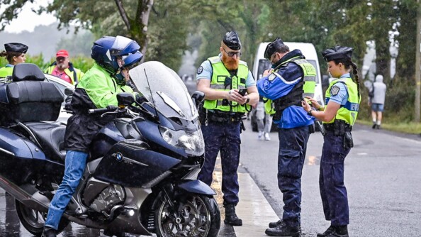 A gauche, une moto et son pilote, à droite, trois gendarmes dont deux effectuent des recherches sur leurs téléphone. Ils sont à un stop de carrefour et il pleut