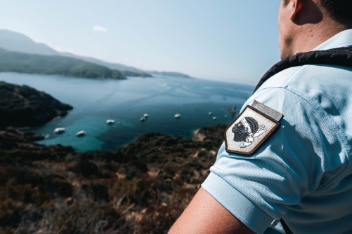 Un gendarme, dont on aperçoit uniquement la manche du polo bleu ciel avec un écusson sur lequel apparait l'emblème du drapeau corse, se tient face à la mer.