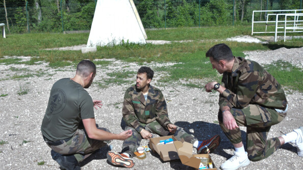 Un instructeur de l'école, en bas de treillis et tee-shirt kaki, est accroupi et discute avec un volleyeur en treillis, assis sur les graviers, les jambes écartées, sa ration de combat ouverte devant lui. A droite, un autre volleyeur, en treillis, un genou au sol, lui aussi devant sa ration de combat ouverte.