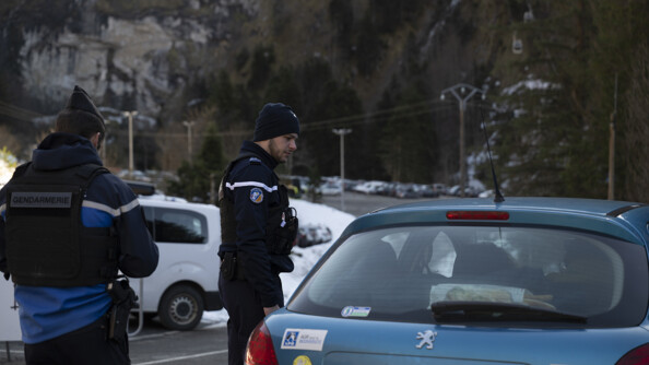 Deux gendarmes, celui de gauche de dos, celui au centre de profil, abordent un véhicule peugeot bleu ciel dans une route à lacets aux pieds de montagnes