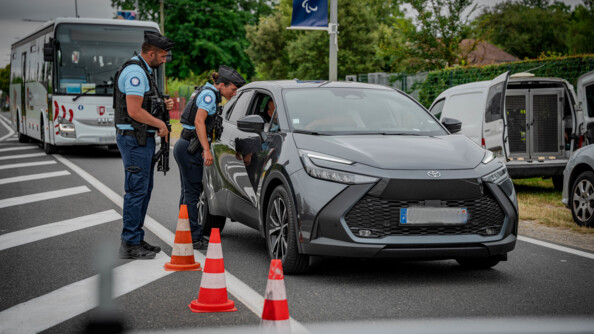 Deux gendarmes, une femme et un homme, de profil, à côté d'un véhicule arrêté sur la voie, à côté de trois plots. La gendarme s'adresse au conducteur par la vitre passager ouverte. Derrière elle, son camarade porte une HK. Derrière la voiture gris foncé, un bus blanc attend. A droite de l'image, l'arrière d'un véhicule cynophile..