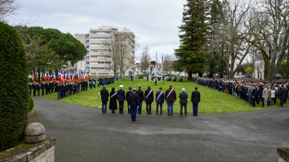 Les autorités au garde à vous, vues de dos, devant la place d'arme en herbe. A gauche, les porte-drapeaux et les troupes et à droite un parterre de personnes civiles.