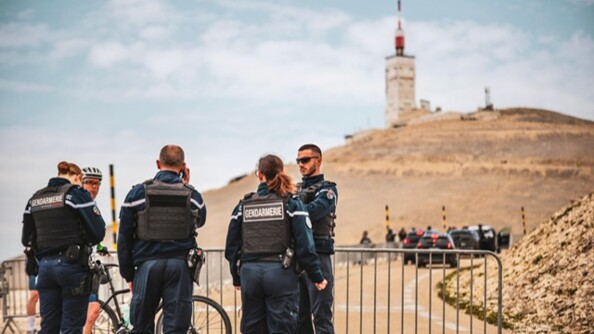 Trois gendarmes de dos, dont un au téléphone, en train d'avancer vers un quatrième de trois-quart face posté devant une barrière en travers de la route conduisant vers le sommet du Mont Ventoux. Un cycliste pied à terre est également devant la barrière et regarde les gendarmes.