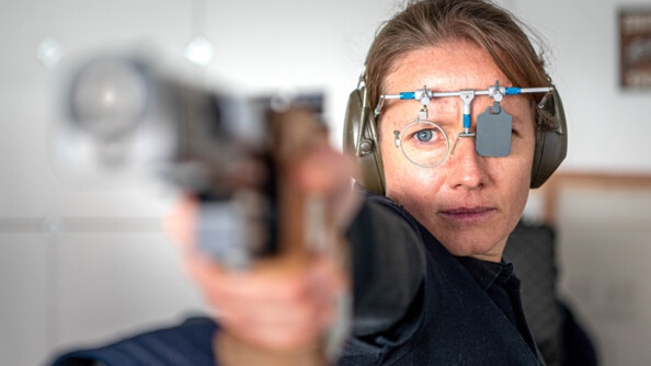 Photo de Gaëlle Edon. On voit son visage, sur ses oreilles se trouve un casque et sur ces yeux, un cache pour obscurcir un de ses yeux. En amorce de l'image, en flou, on voit sa main et son arme qu'elle pointe devant elle, déterminée.