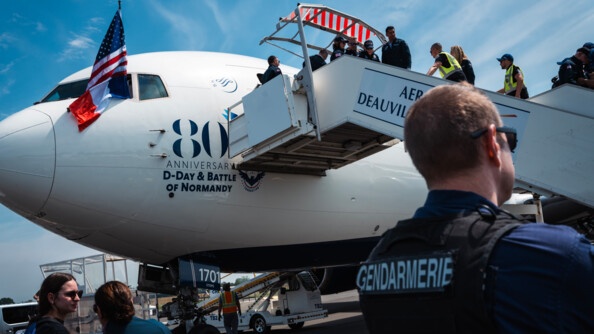 Vue en contre-plongée sur la descente d'escalier à la sortie d'un avion, d'un vétéran. Entouré de persones l'aidant (gendarmes, américains). l'avion est blanc et siglé aux 80 ans du débarquement. Au premier plan tout à droit, buste de dos d'un gendarme