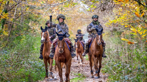 Quatre cavaliers de la Garde républicaine en treillis et armés de Famas traversent les bois sur leurs montures, sur une piste forestière.