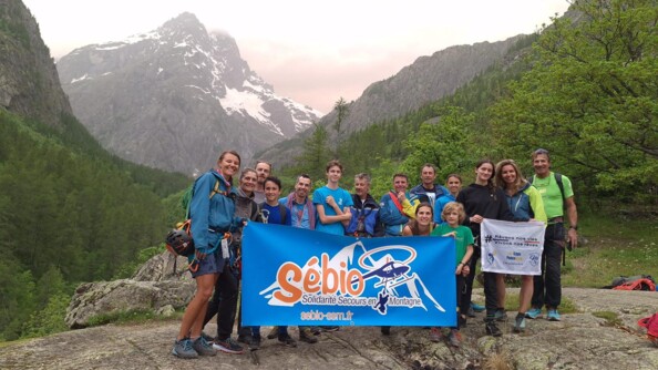 Quinze personnes tenant une banderolle "Sébio Solidarité Secours en Montagne". Derrière eux, se trouve des montagnes et des forêts.