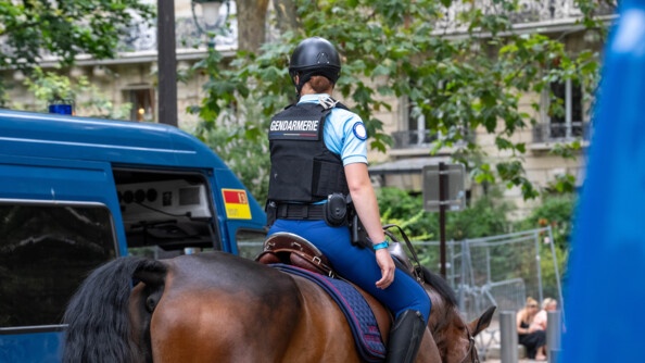 Une cavalière évolue sur son cheval marron, dans les rues de Paris. On la voit de dos, portant un gilet pare-balles, sur lequel est inscrit Gendarmerie. On aperçoit un camion bleu de la gendarmerie garé à sa gauche, et dont la porte est ouverte.