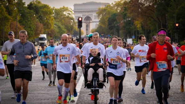 Des coureurs, vêtus d'un T-shirt blanc avec un numéro sur le ventre, courent sur une avenue pavée, en poussant le fauteuil roulant d'une jeune femme blonde, souriante, portant des tresses. On aperçoit l'arc de triomphe en arrière plan.