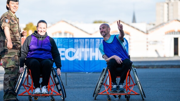 Deux personnes dans des fauteuils roulants rouge, devant une grille surmontée d'un panneau décathlon. l'homme de droite fait le sign eV de sa main gauche. A gauche de l'image, une gendarme en treillis sourit