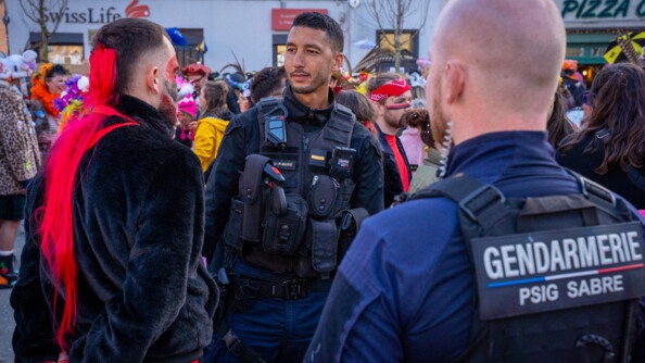 La photo repr&eacute;sente un gendarme, debout dans la rue aux c&ocirc;t&eacute;s des carnavaleux, qui portent un d&eacute;guisement, lors du carnaval de Bergues.